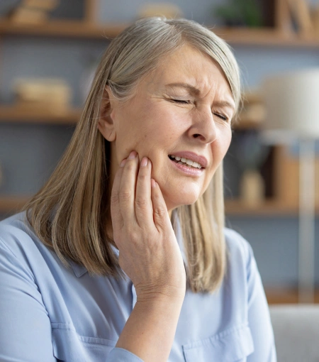 Middle-aged woman touching her cheek in pain, dental office setting with shelves in background.