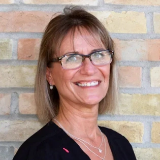 Woman wearing glasses smiling at camera against brick wall background, dental office portrait.
