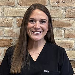 Young woman smiling at camera wearing black scrubs against brick wall background.