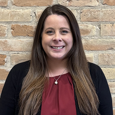 Professional headshot of smiling woman with long brown hair against brick wall background.